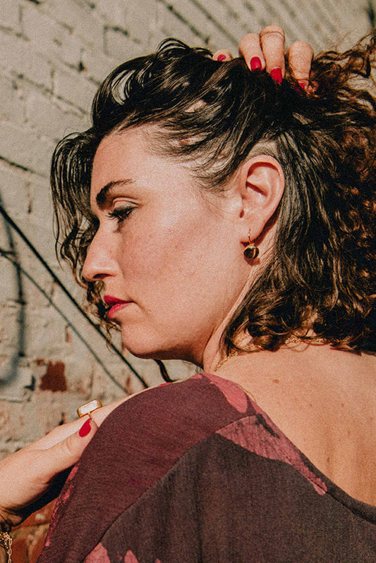 Model wearing Hand printed top, gold chain, drop earrings against a weathered brick wall. 