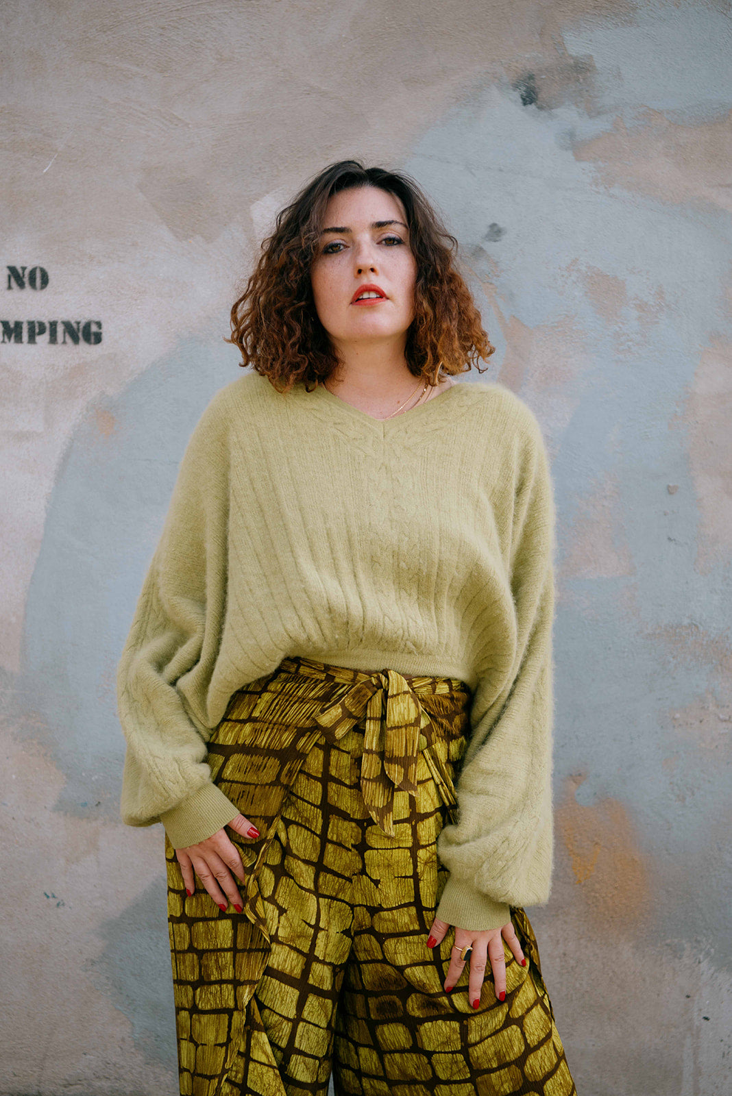Woman wearing a light green sweater and patterned pants against a textured wall.