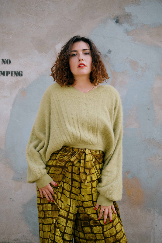 Woman wearing a light green sweater and patterned pants against a textured wall.