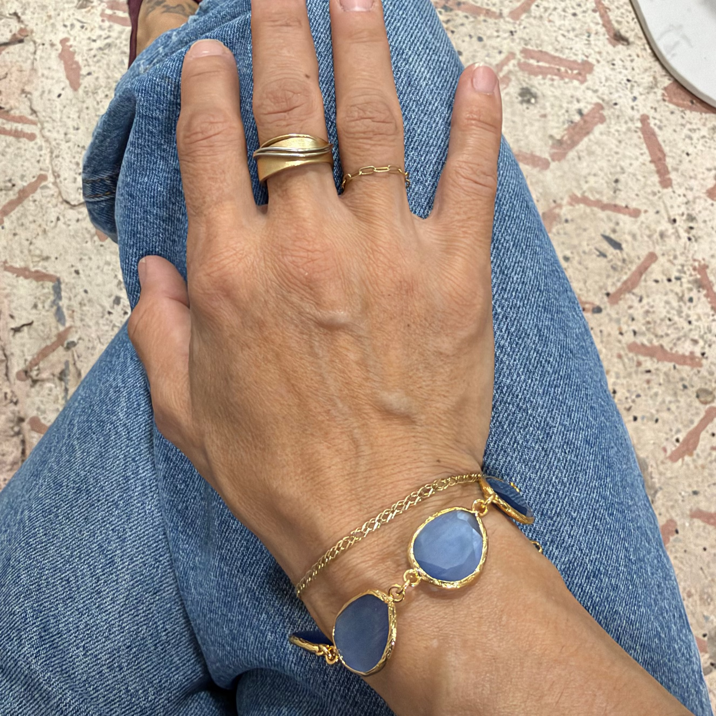 Hand wearing gold bracelets and rings on a denim surface with a textured floor background